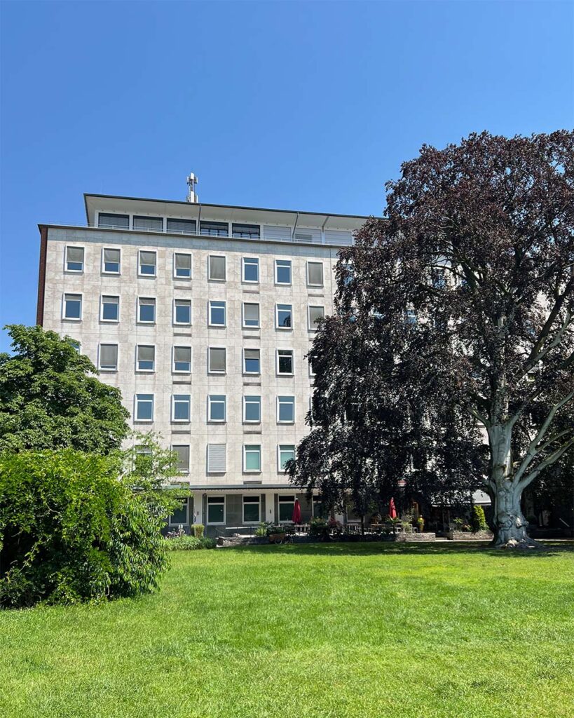 Außenansicht des EFTAS-Firmengebäudes, ein mehrstöckiger heller Bürobau mit angrenzender Grünfläche, Bäumen und Außenterrasse unter blauem Himmel.