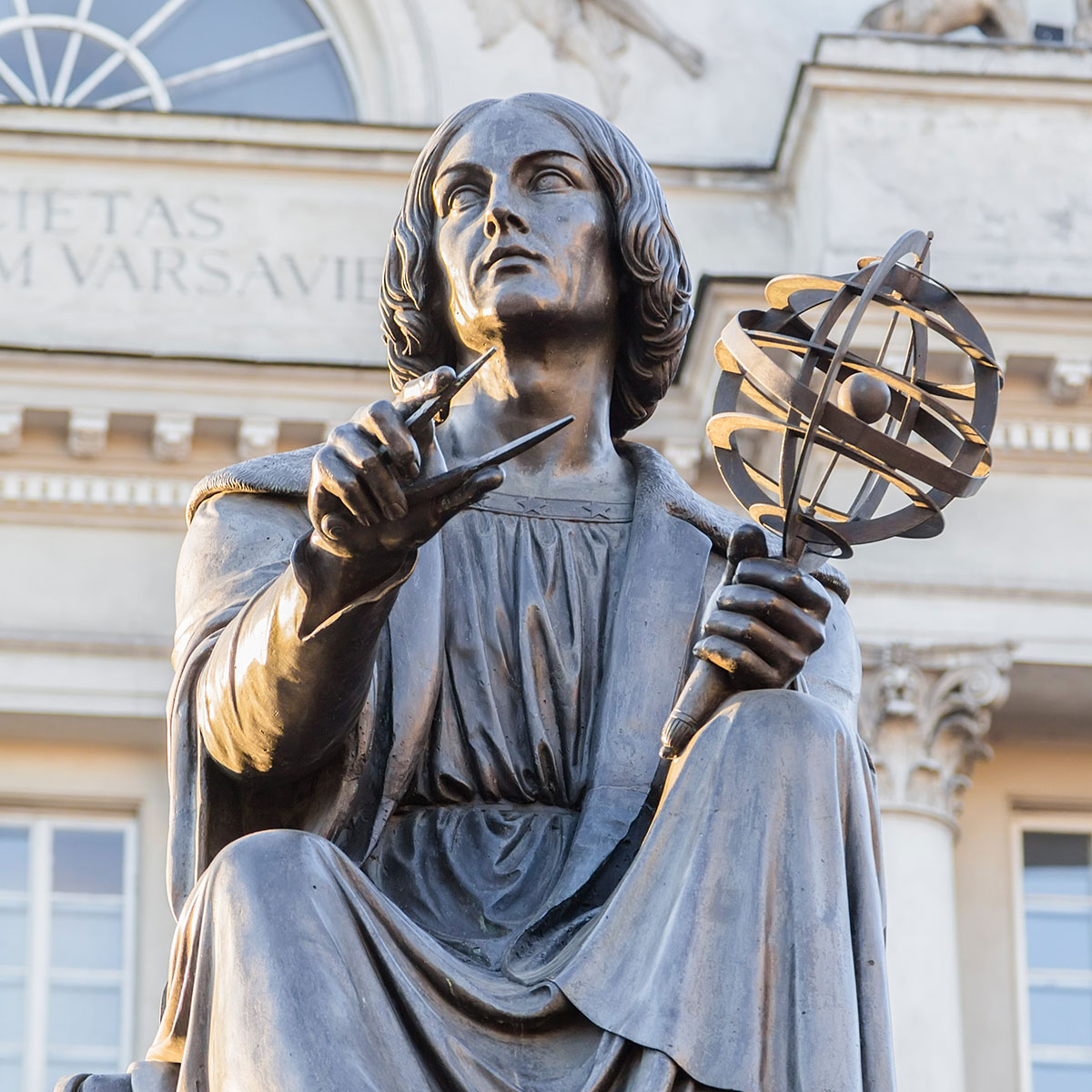 Bronzestatue eines Gelehrten mit Zirkel und Himmelsglobus vor einer historischen Gebäudefassade.