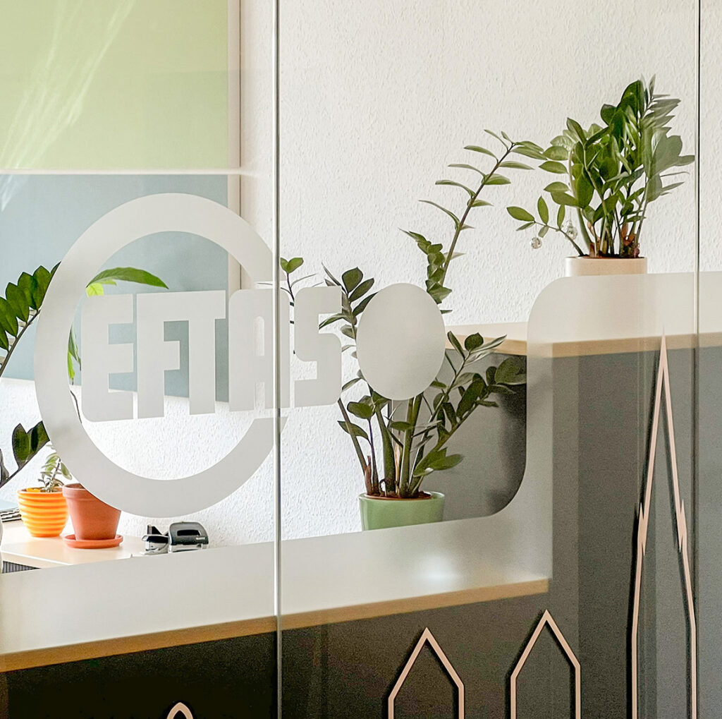 Empfangsbereich von EFTAS mit dem Firmenlogo auf einer Glaswand, Zimmerpflanzen und einer stilisierten Stadtsilhouette.